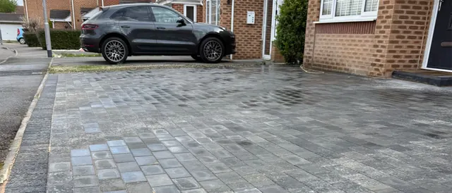 Wide grey block paving driveway in mixed tones with a charcoal edging course, in front of a brick house