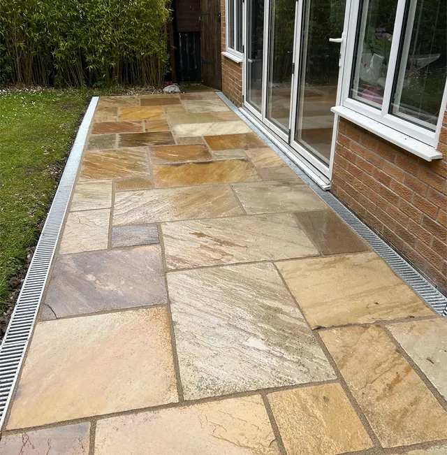 Golden Indian sandstone patio beside patio doors, with a drainage channel along the house wall